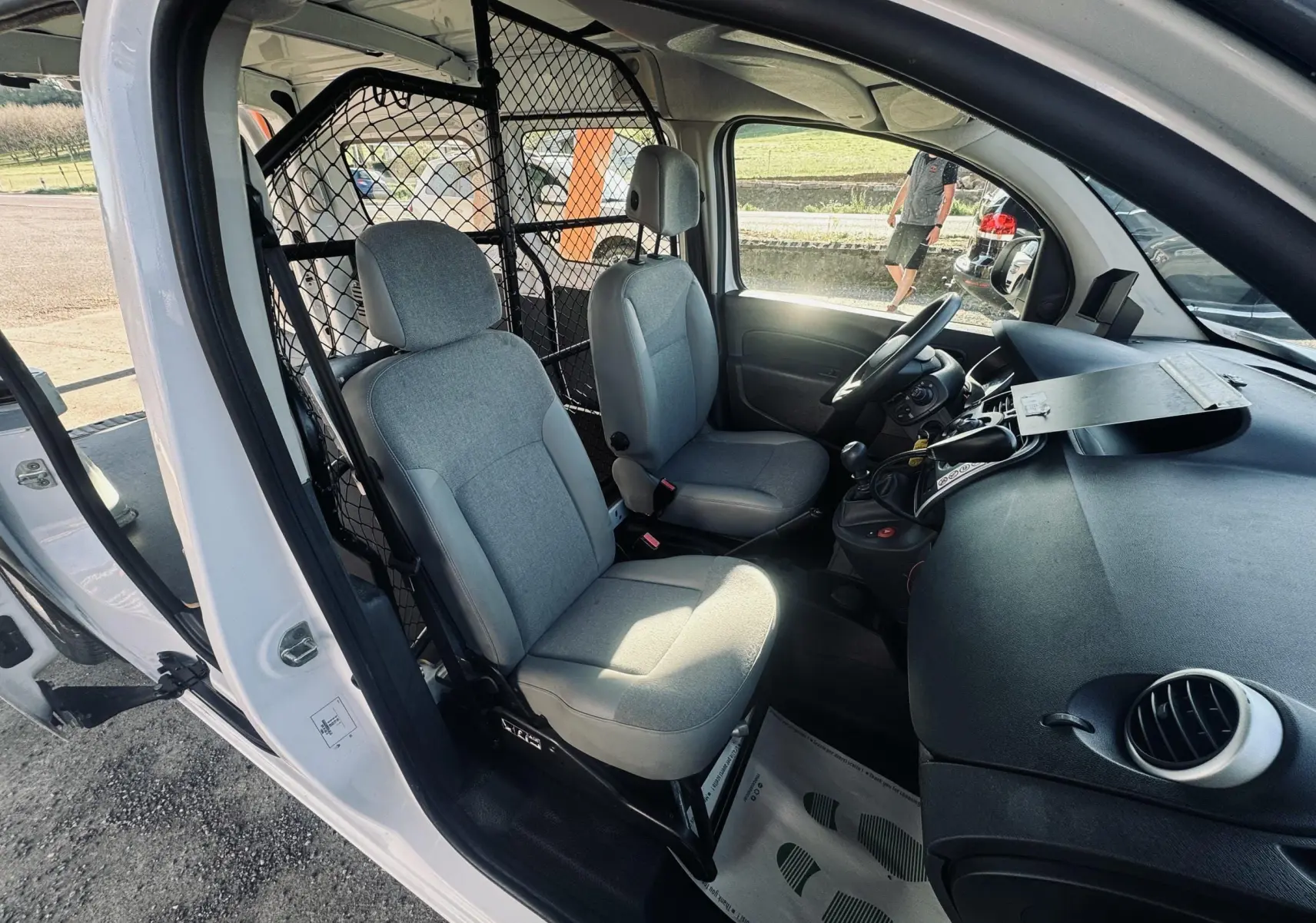 Vue intérieure côté conducteur du Renault Kangoo blanc, montrant les sièges gris et la cloison de séparation métallique.