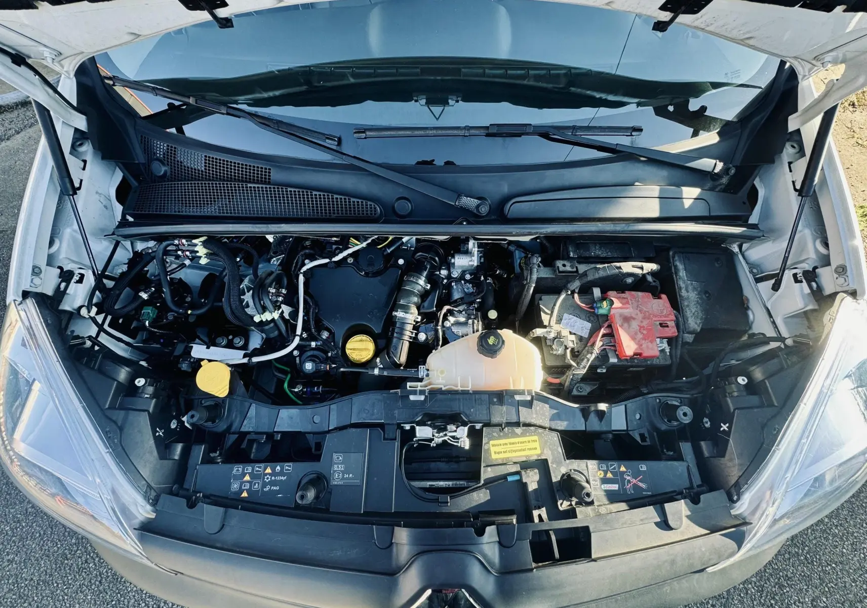 Vue plongeante sur le moteur d’un Renault Kangoo blanc avec capot ouvert, montrant les composants et la batterie.