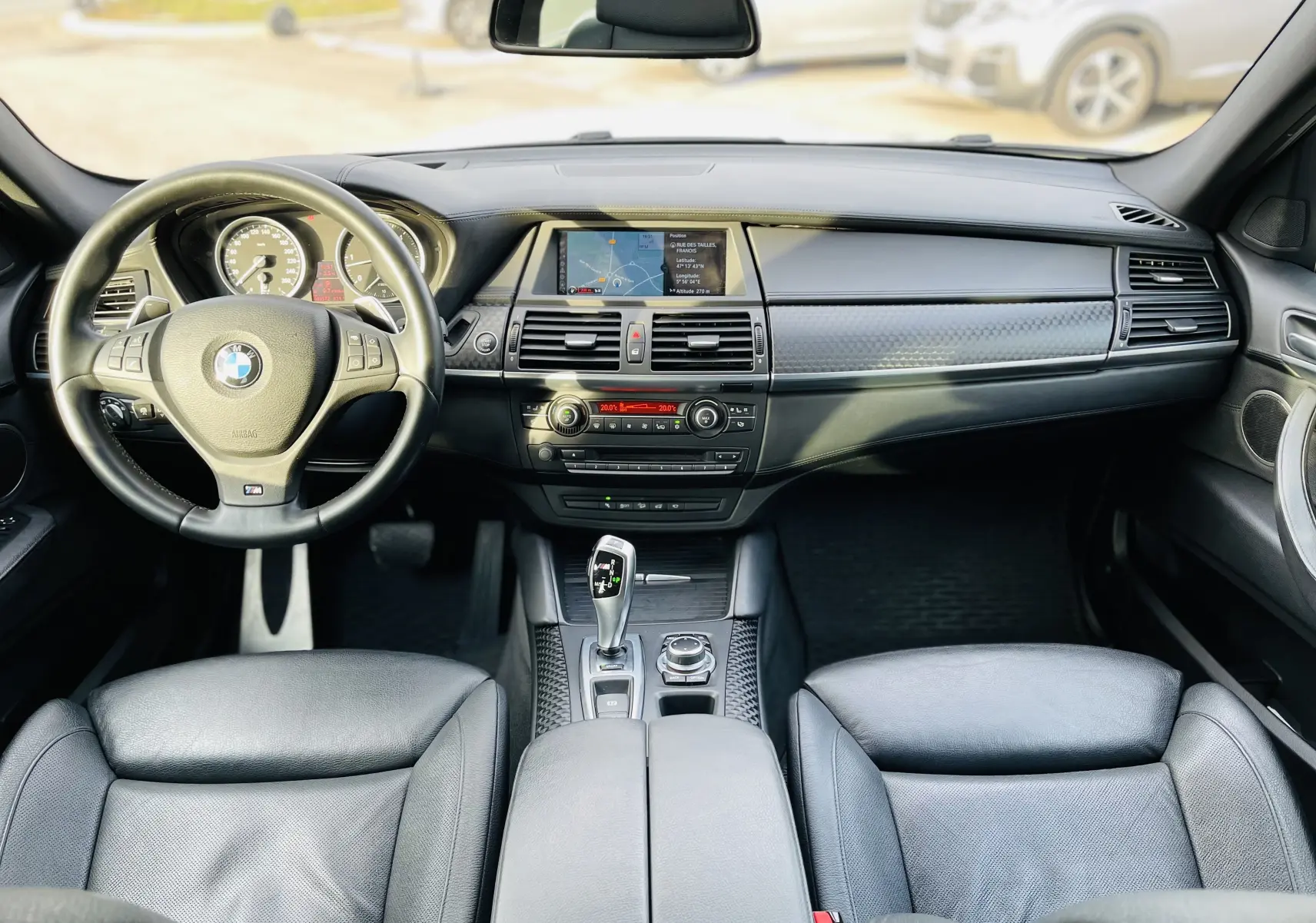 Vue intérieure avant du BMW X6 50D blanc, tableau de bord noir avec écran central et volant M cuir.