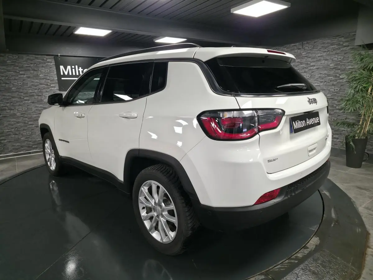 Vue 3/4 arrière droite d'un Jeep Compass blanc avec vitres surteintées et jantes alliage 18 pouces en intérieur showroom.