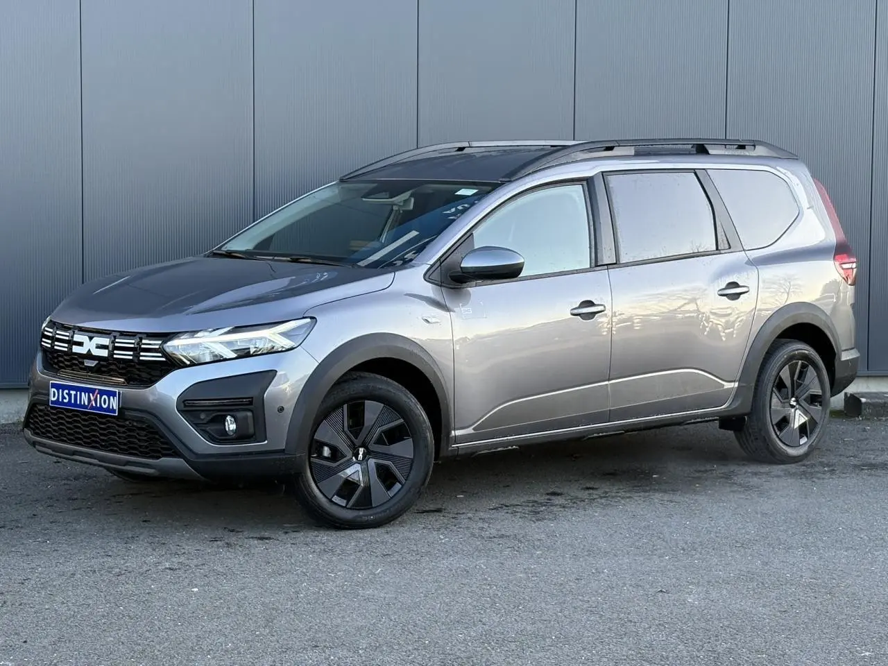 Vue 3/4 avant droite du Dacia Jogger gris schiste avec barres de toit et jantes noires distinctives.