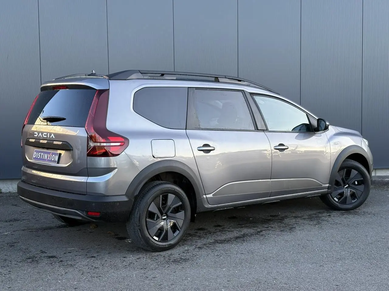 Dacia Jogger gris schiste en 3/4 arrière droit, avec barres de toit et jantes noires distinctives.