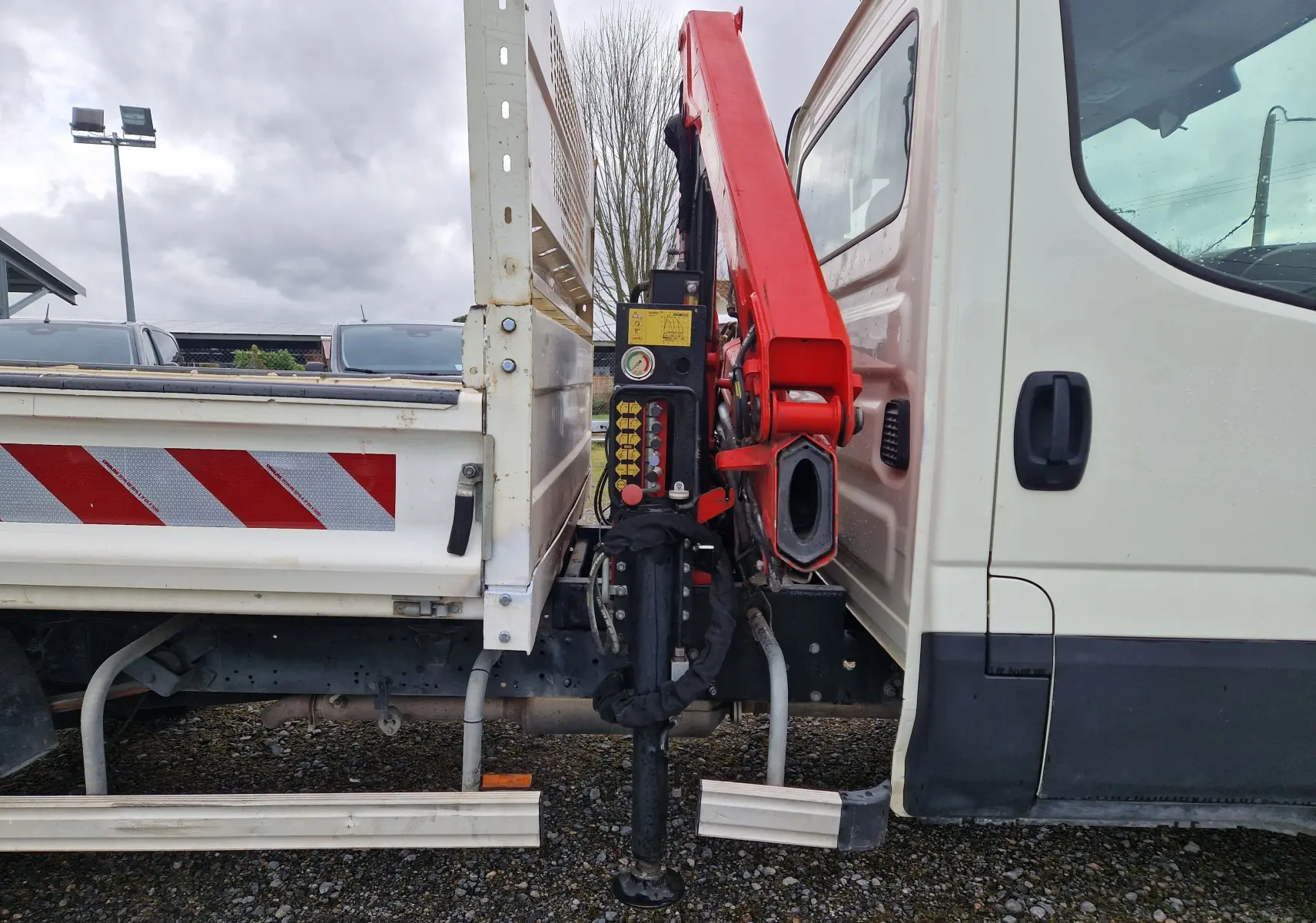 Vue latérale droite d’un Iveco Daily blanc avec benne et grue rouge Palfinger déployée entre cabine et plateau.