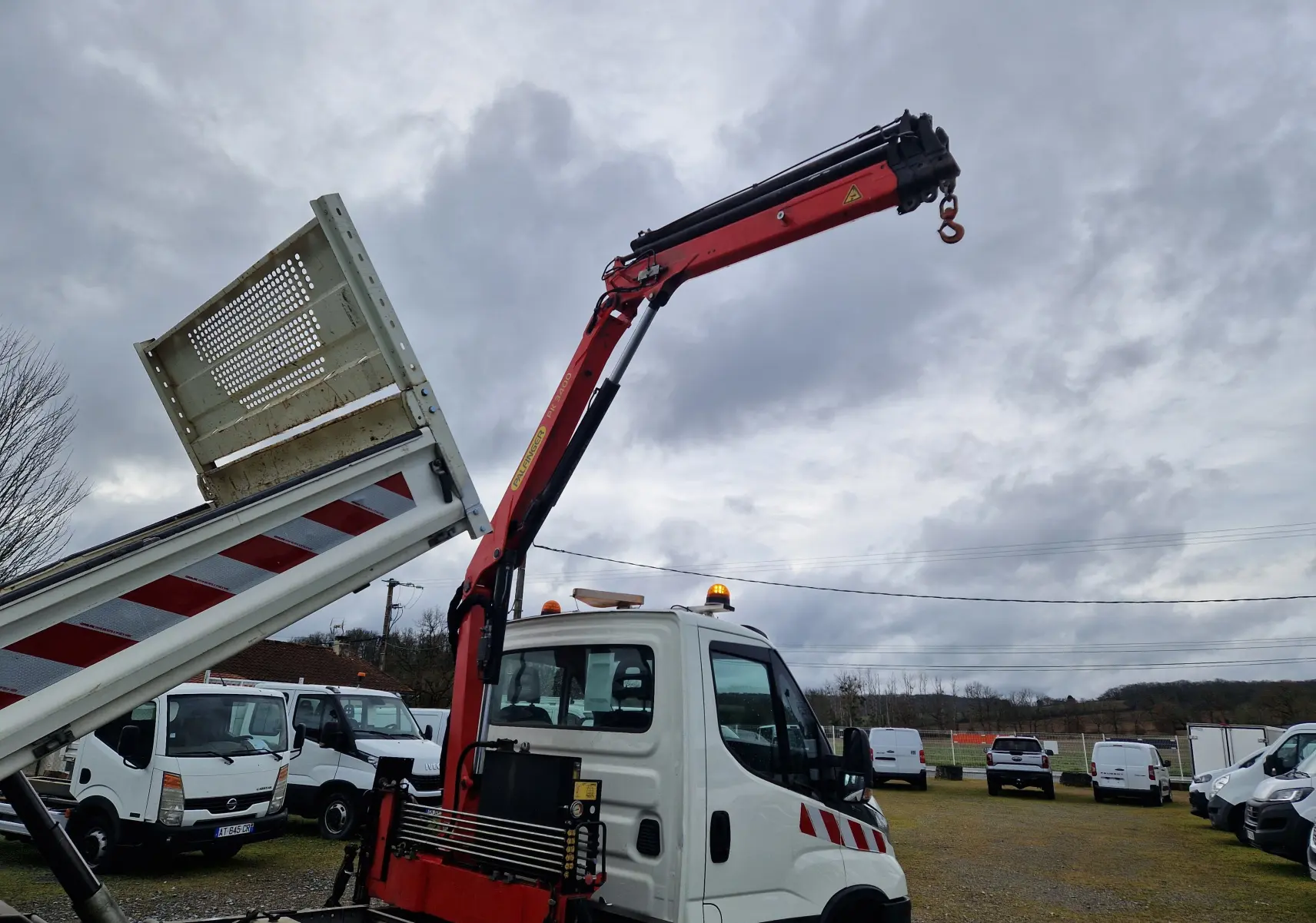 Vue 3/4 arrière droite d’un Iveco Daily blanc avec benne relevée et grue Palfinger rouge déployée.
