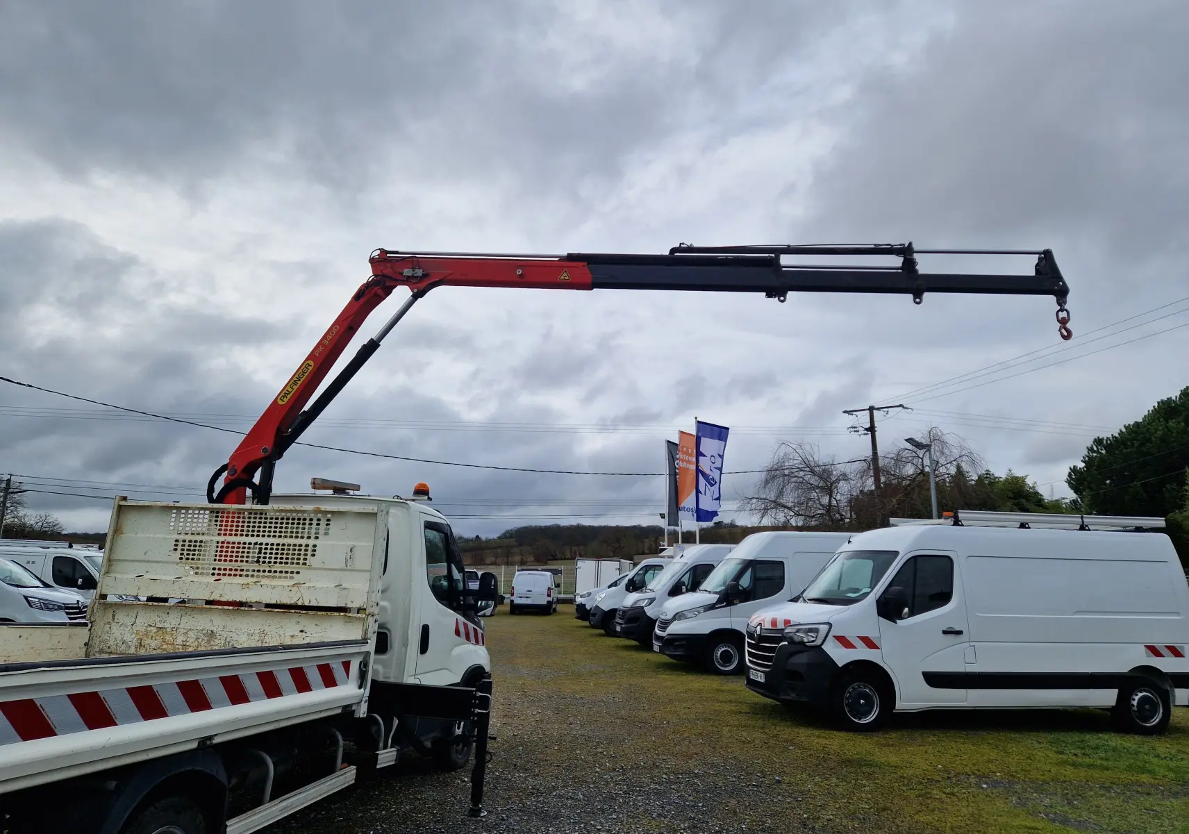 Vue 3/4 arrière gauche d’un Iveco Daily blanc avec benne et grue Palfinger déployée en rouge et noir.