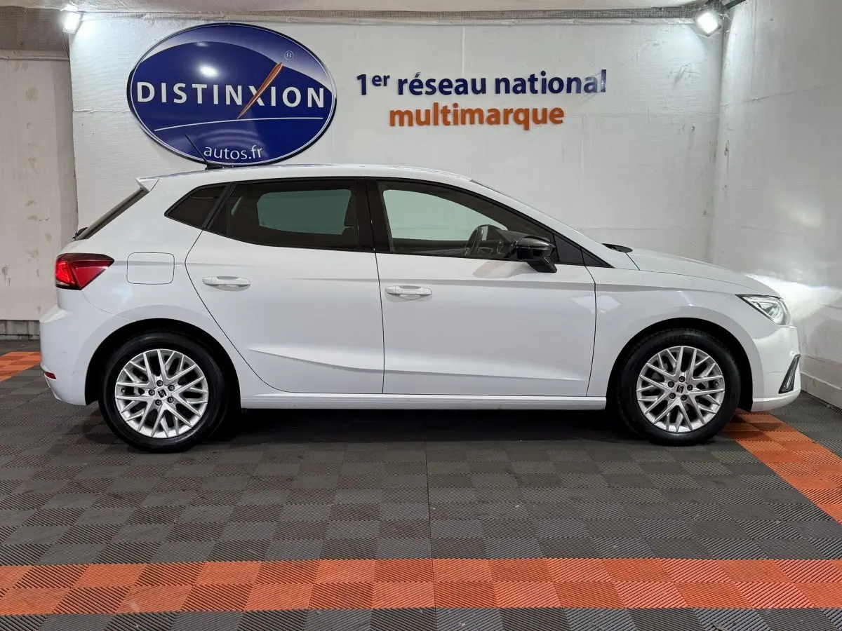 Vue de profil côté gauche d'une SEAT Ibiza 1.0 TSI 110 FR blanche avec jantes alliage multibranches en intérieur showroom.