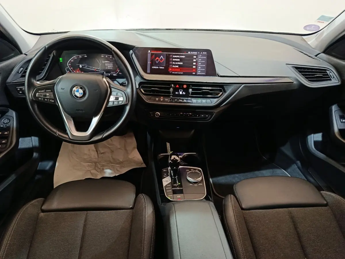 Vue intérieure avant de la BMW Série 1 118i Edition Sport 2023, tableau de bord noir avec écran tactile et volant cuir multifonctions.