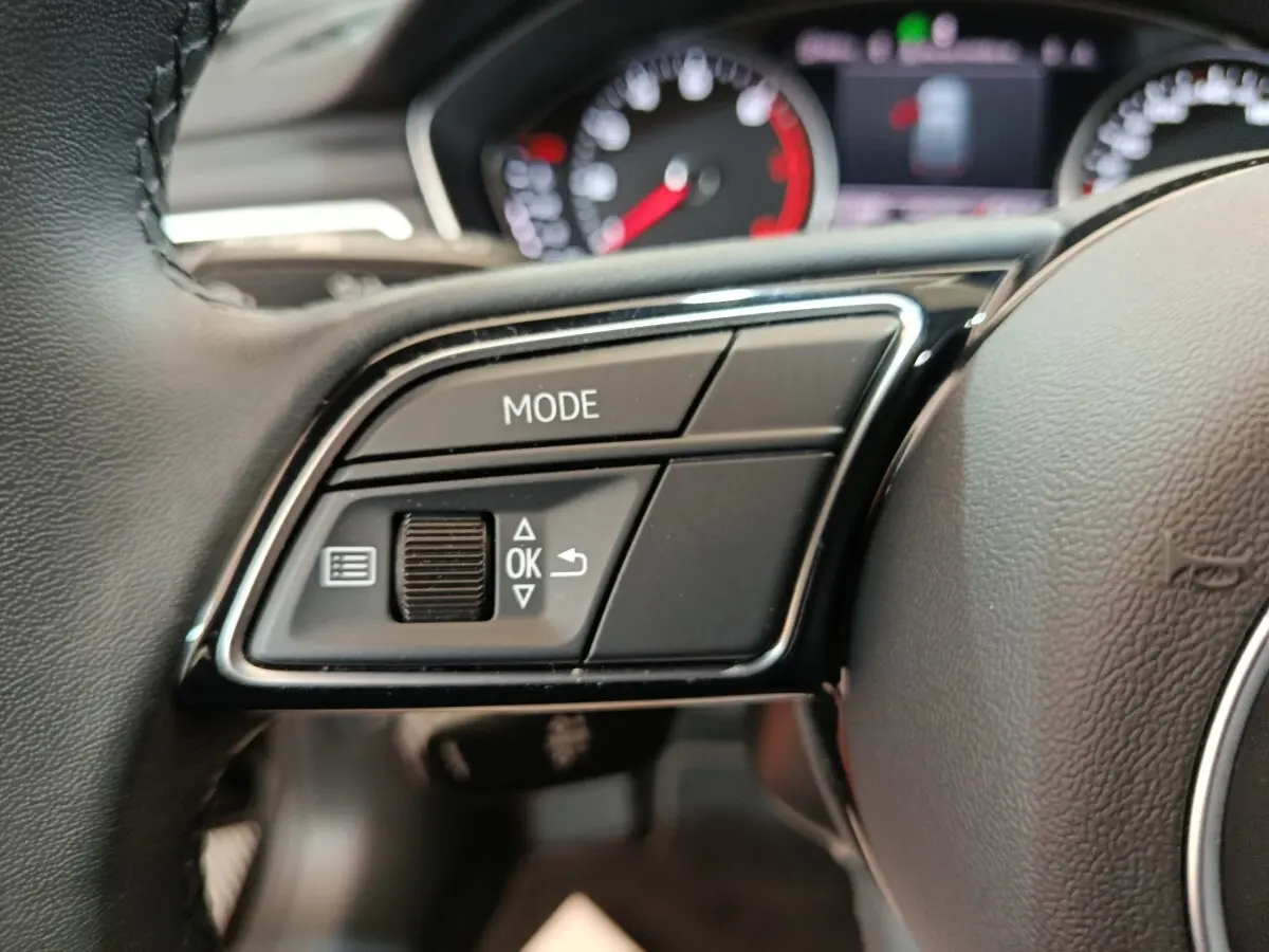 Gros plan sur les commandes gauche du volant cuir noir de l’Audi A4 35 TFSI 2024 avec tableau de bord flou en arrière-plan.