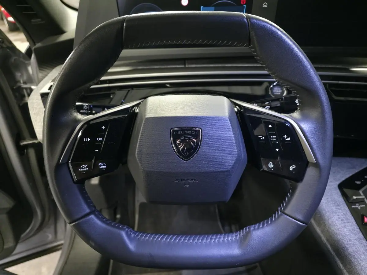 Volant cuir noir avec logo Peugeot en vue frontale, tableau de bord et console centrale tissu gris visibles.