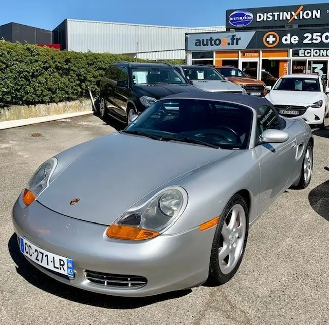 Porsche Boxster 986 gris 3/4 avant droit, capote noire, feux avant ovales et jantes argentées visibles.