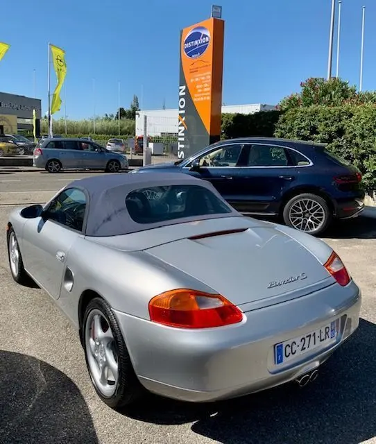 Vue 3/4 arrière d'une Porsche Boxster 986 gris argent avec capote noire, stationnée en extérieur par temps clair.
