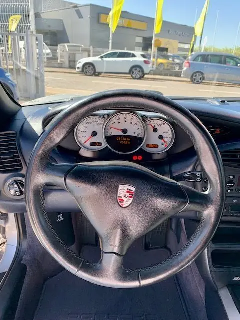 Vue intérieure centrée sur le volant noir avec logo Porsche et tableau de bord blanc d'une Boxster 986 gris 2001.