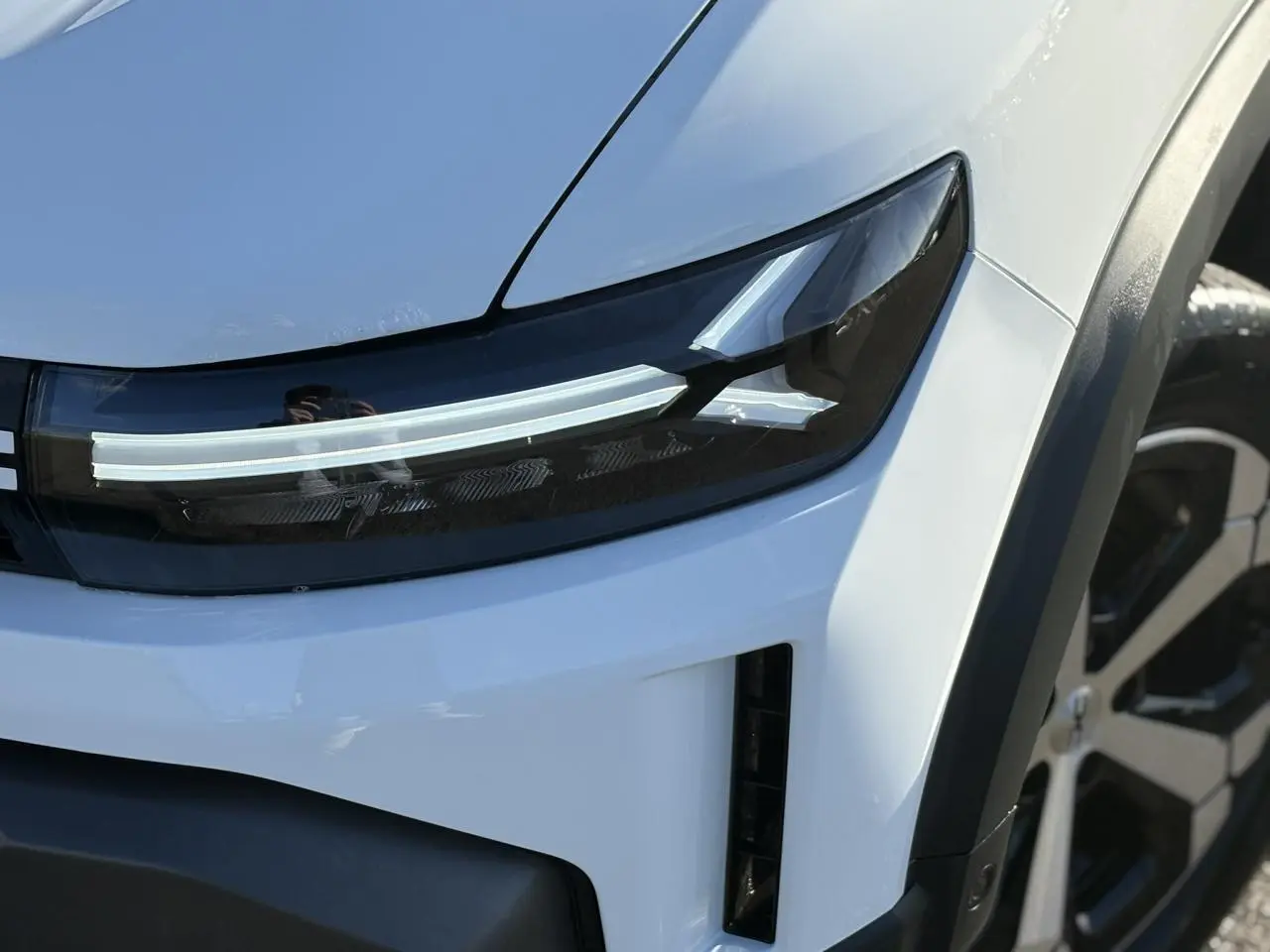 Gros plan sur le phare avant droit blanc glacier du Dacia Duster 2026 avec jante alliage visible en arrière-plan.