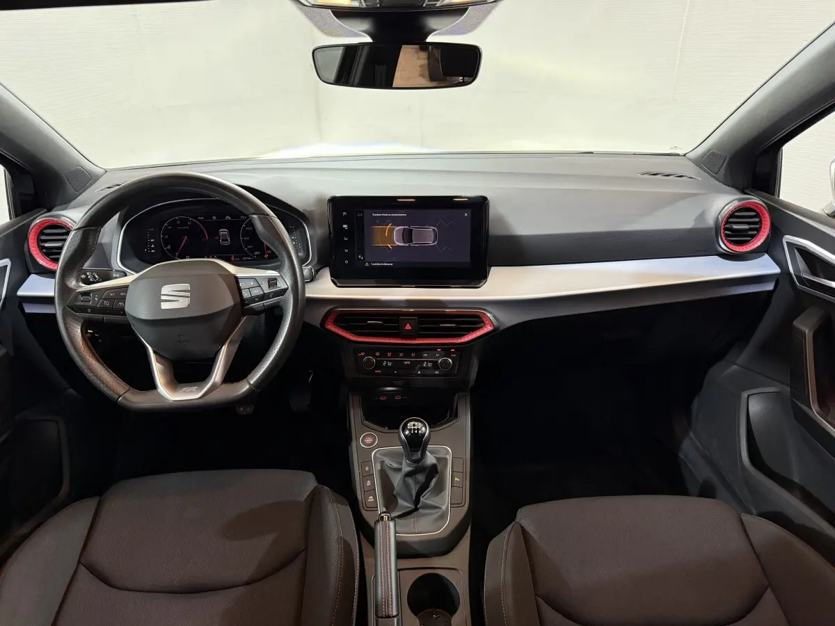 Vue intérieure frontale du tableau de bord de la SEAT Ibiza 2024 avec volant cuir alu et touches rouges autour des aérateurs.