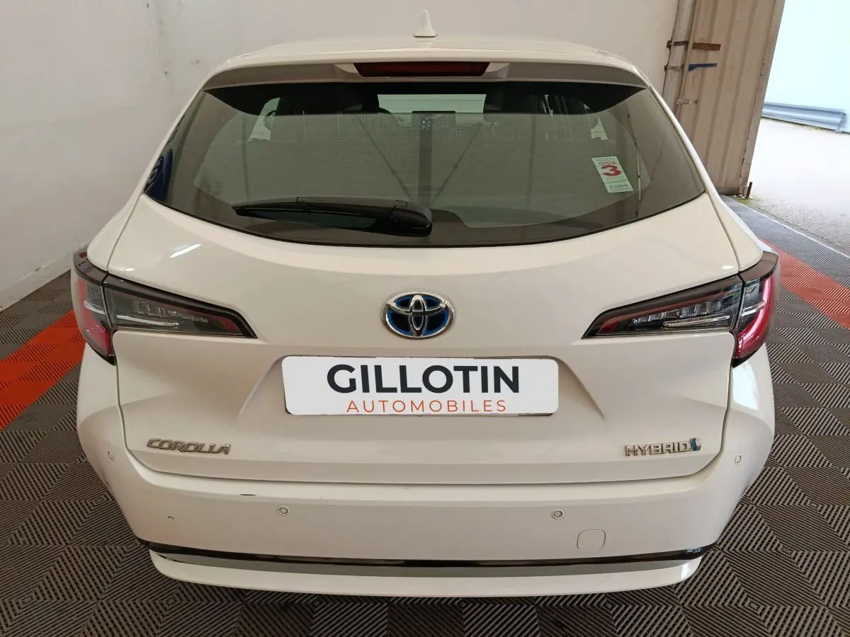 Vue arrière d'une Toyota Corolla Hybride blanche 2020, avec feux LED et logo Toyota visible sur un sol à damier noir et rouge.