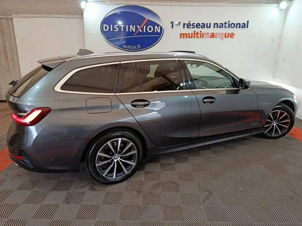 BMW Série 3 Touring gris clair vue de profil côté droit, avec feux arrière allumés dans un showroom.