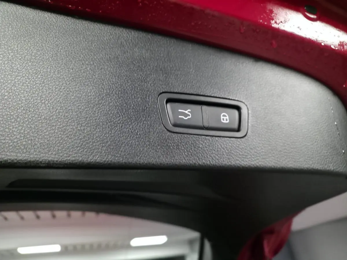 Gros plan sur le bouton de fermeture électrique du hayon, intérieur noir, bord rouge du coffre visible.