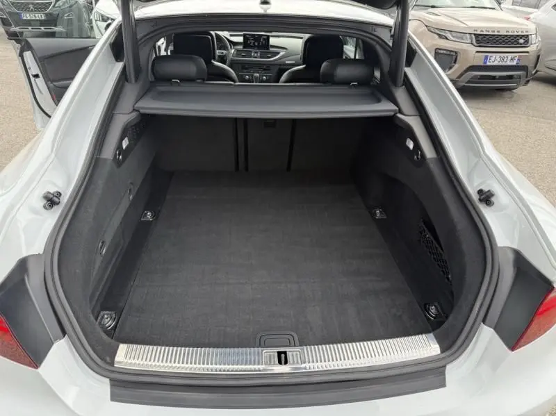 Coffre ouvert vu de l'arrière d'une Audi A7 Sportback blanche, intérieur spacieux avec cache-bagages déployé.