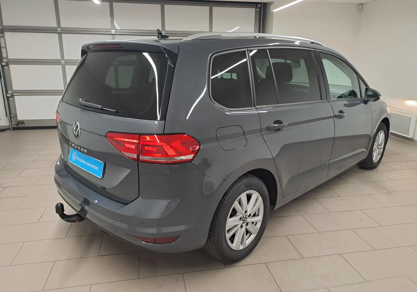 Vue 3/4 arrière droite d'un Volkswagen Touran gris dauphin avec attelage démontable en intérieur showroom.