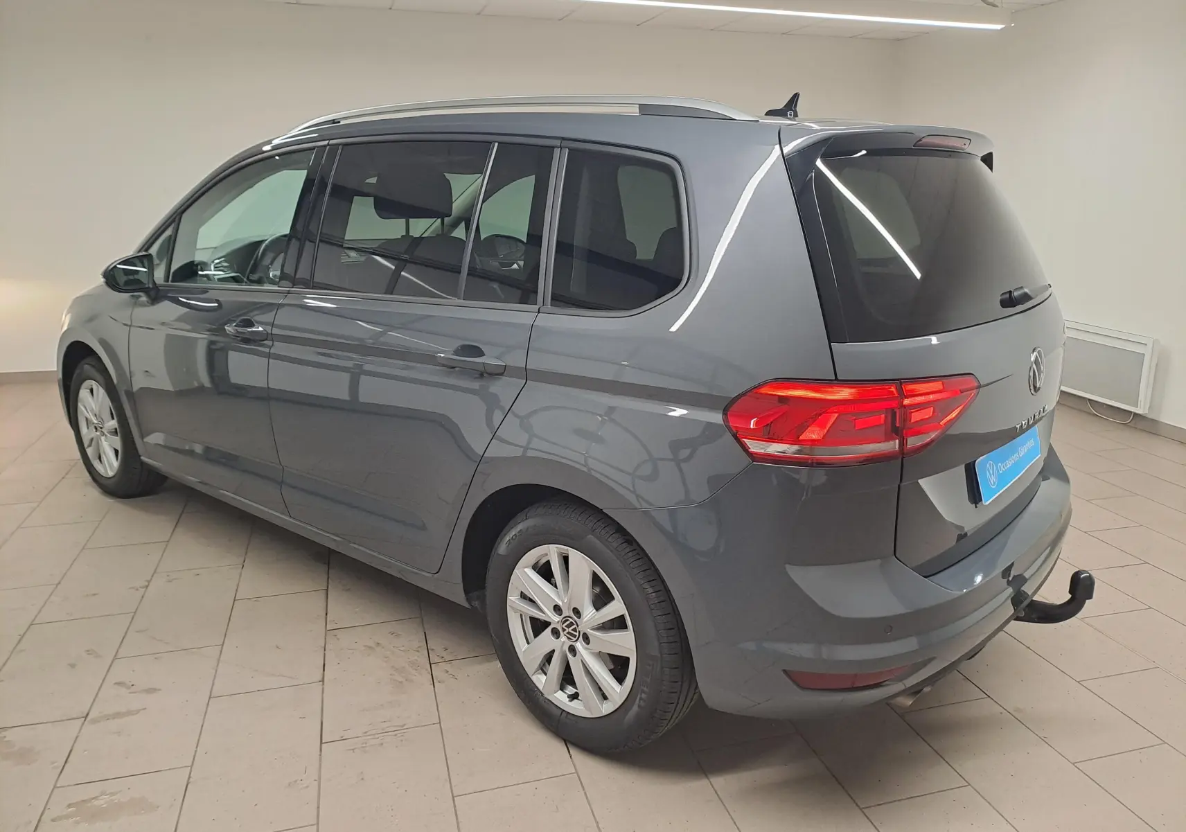 Vue 3/4 arrière droite d'un Volkswagen Touran gris dauphin 2024 avec attelage démontable et vitres teintées.