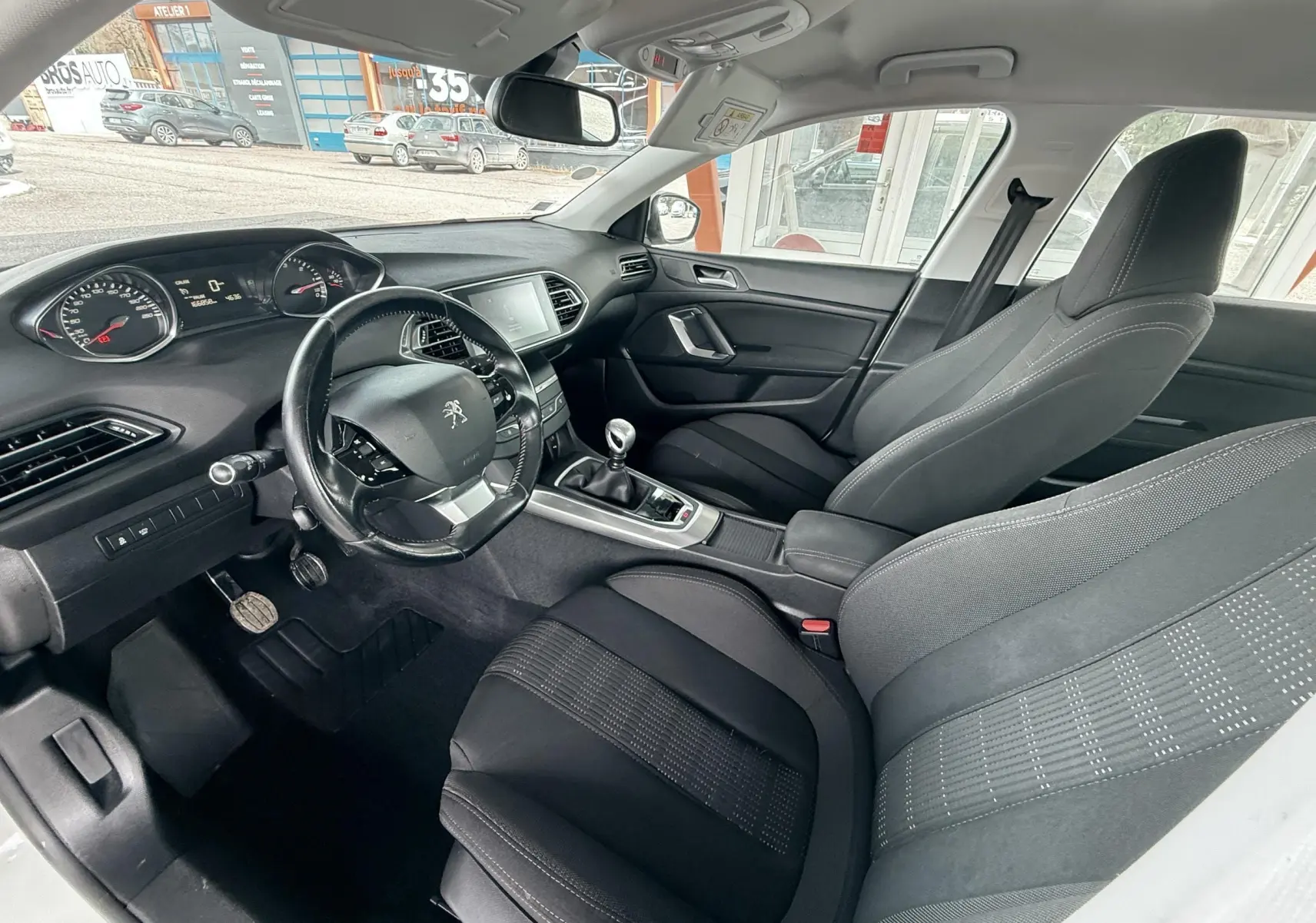 Vue intérieure côté conducteur de la Peugeot 308 blanche, montrant le tableau de bord et les sièges en tissu noir.