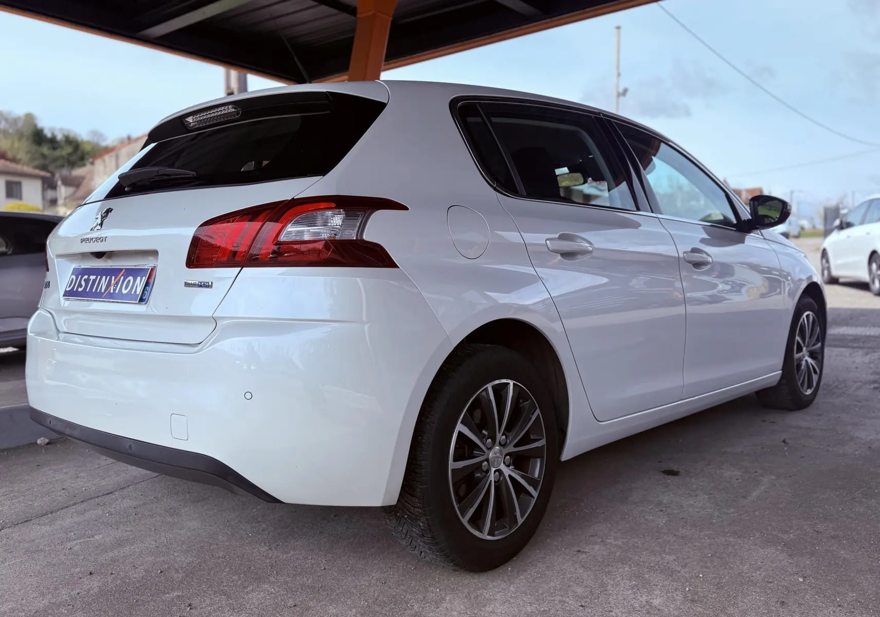 Peugeot 308 blanc nacré vue 3/4 arrière droit sous abri, avec feux arrière rouges et jantes alliage foncé.