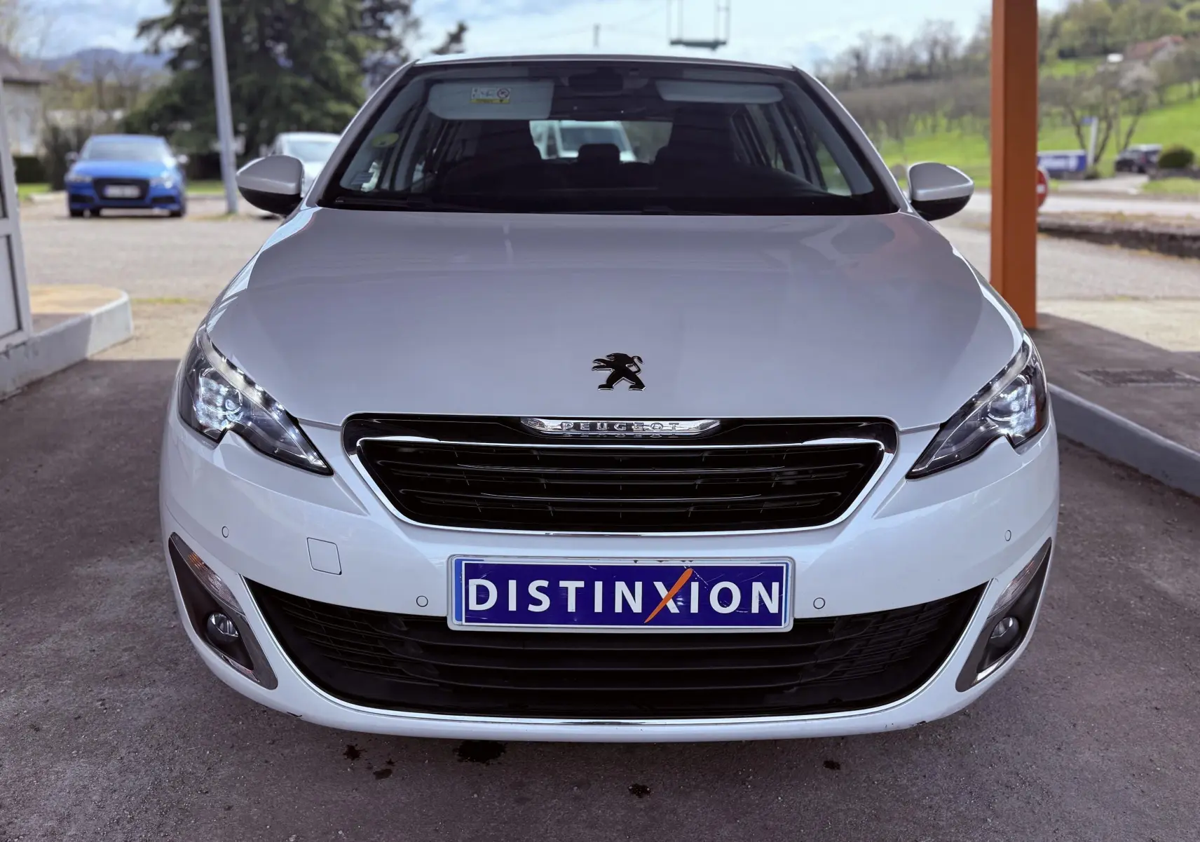 Vue frontale d'une Peugeot 308 blanche nacrée avec phares allumés et plaque d'immatriculation Distinxion.