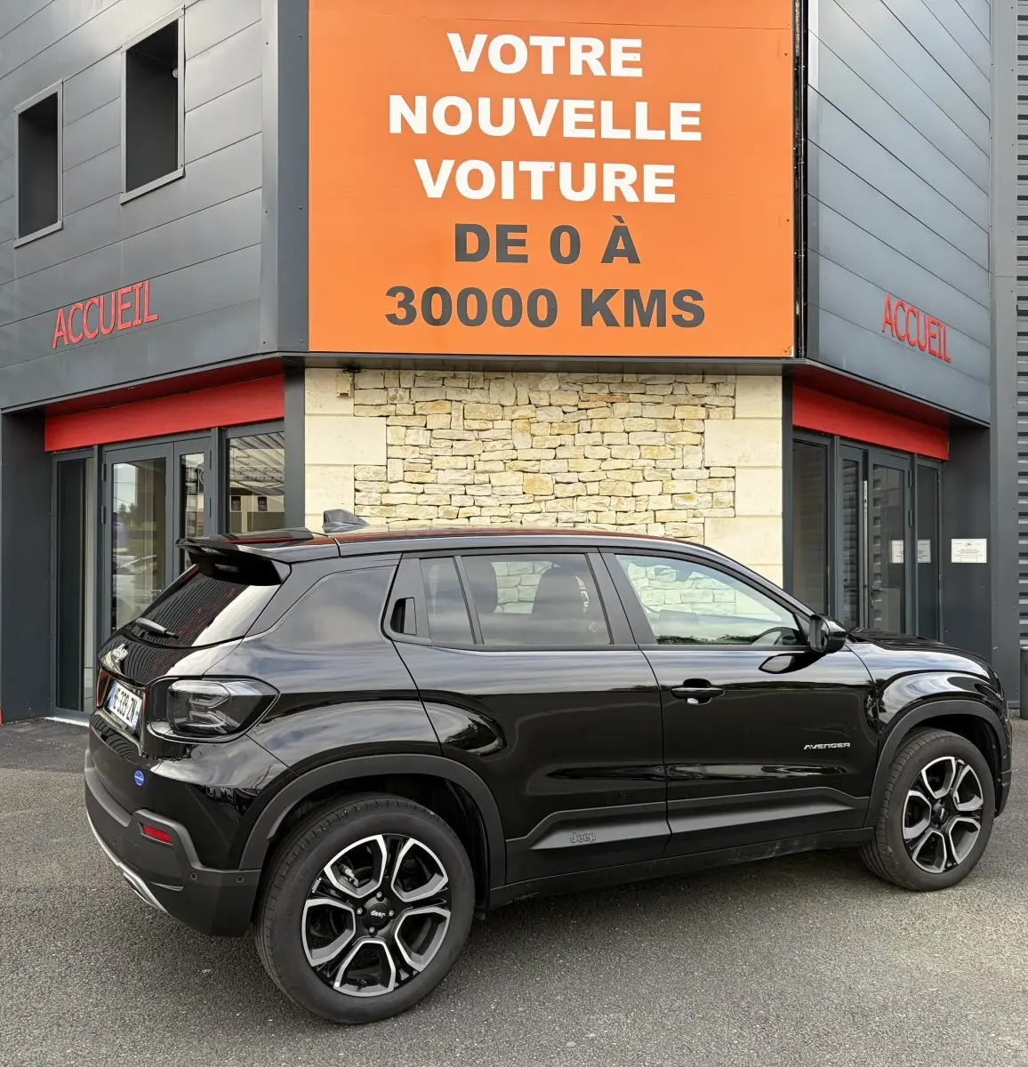 Jeep Avenger noir en vue de profil côté droit, garé devant un bâtiment avec panneau orange "Votre nouvelle voiture".