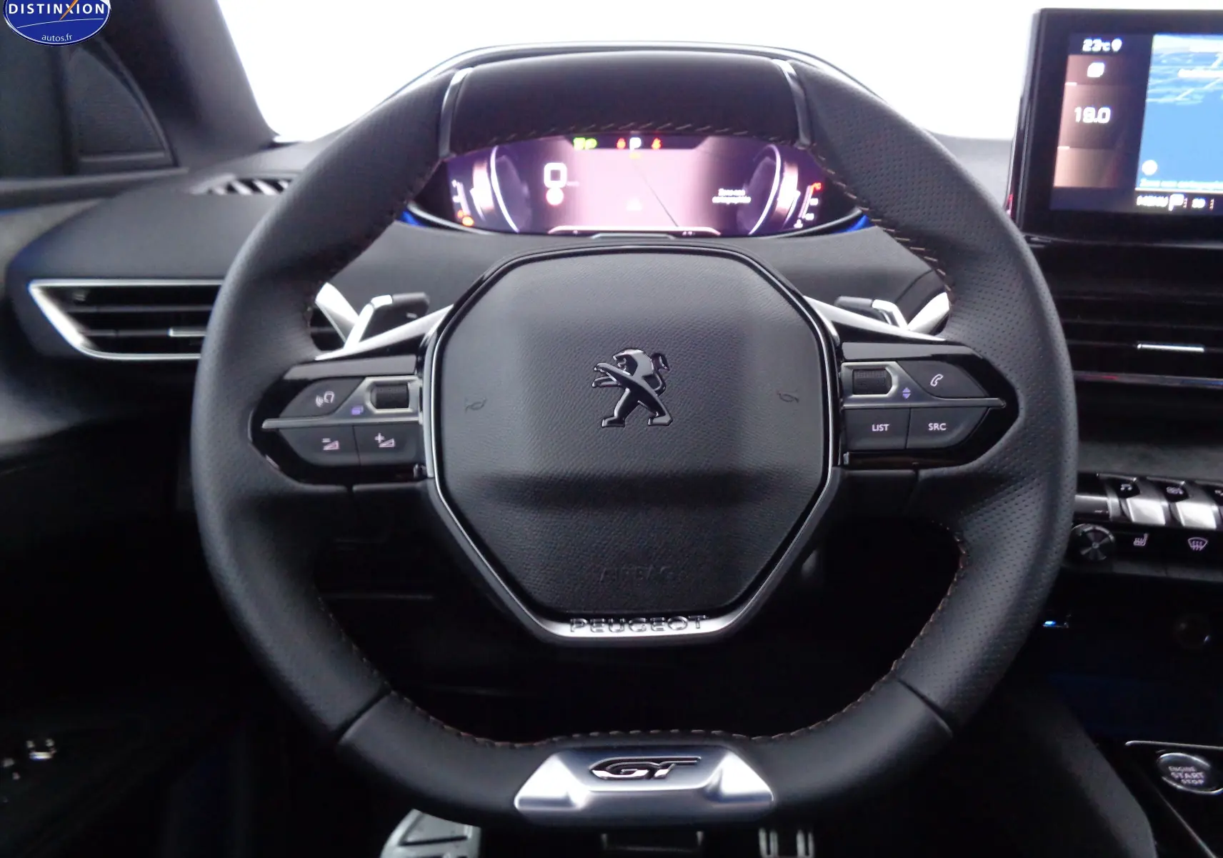 Volant cuir noir au design compact avec logo Peugeot et badge GT, tableau de bord numérique en arrière-plan.