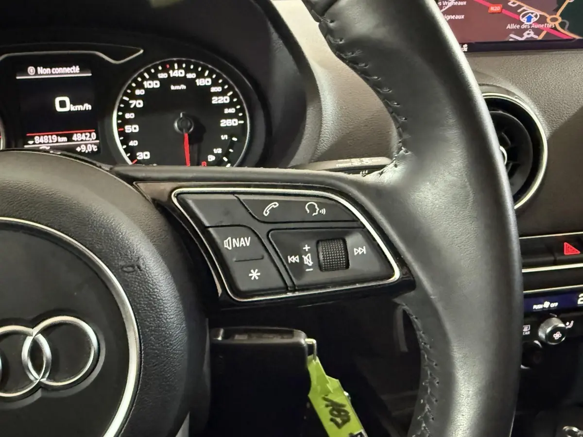 Vue rapprochée du volant cuir noir d’Audi A3 2018 avec commandes audio et tableau de bord digital.