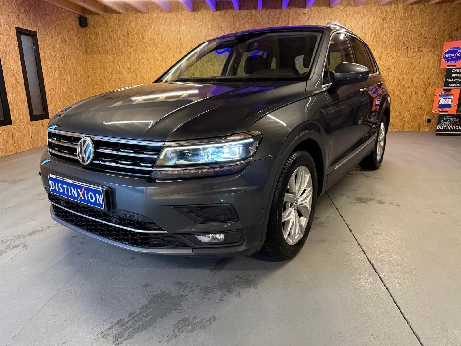 Volkswagen Tiguan gris 3/4 avant droit avec phares allumés dans un garage à murs en bois.