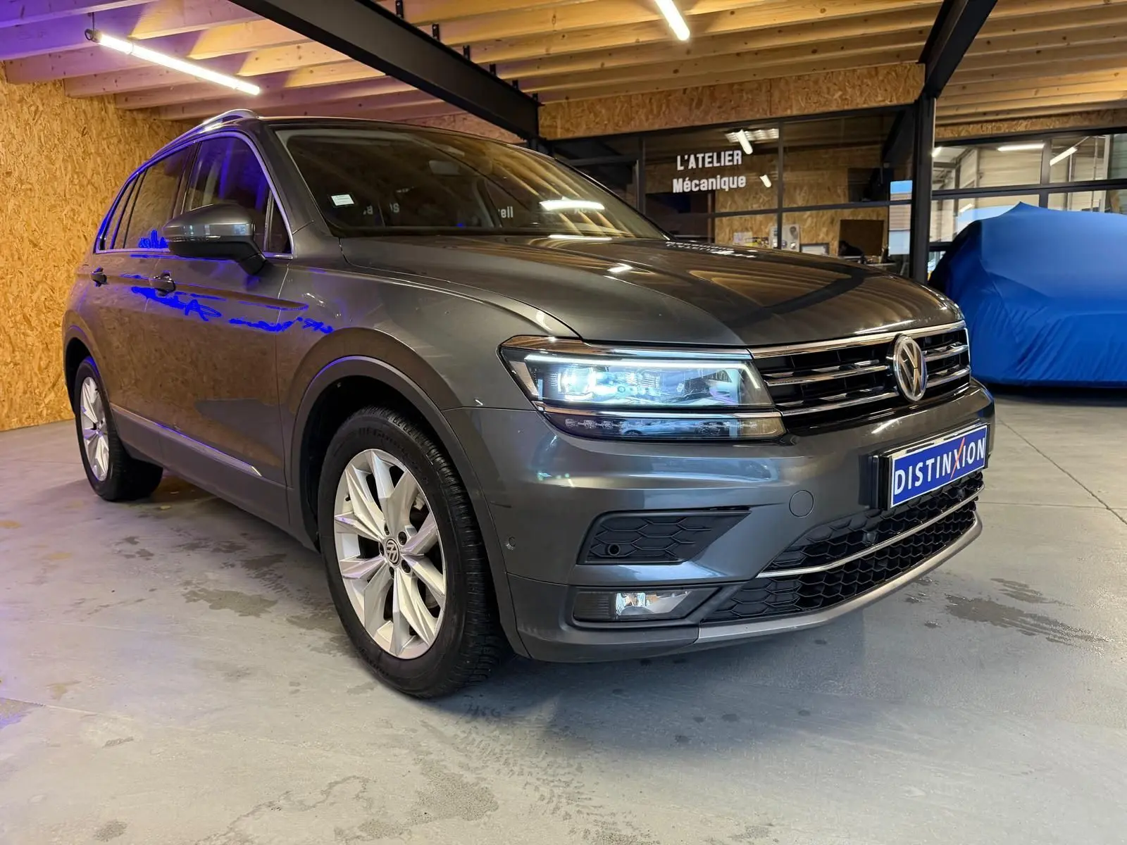 Volkswagen Tiguan gris 3/4 avant droit, phares allumés, jantes alu, dans un garage intérieur.
