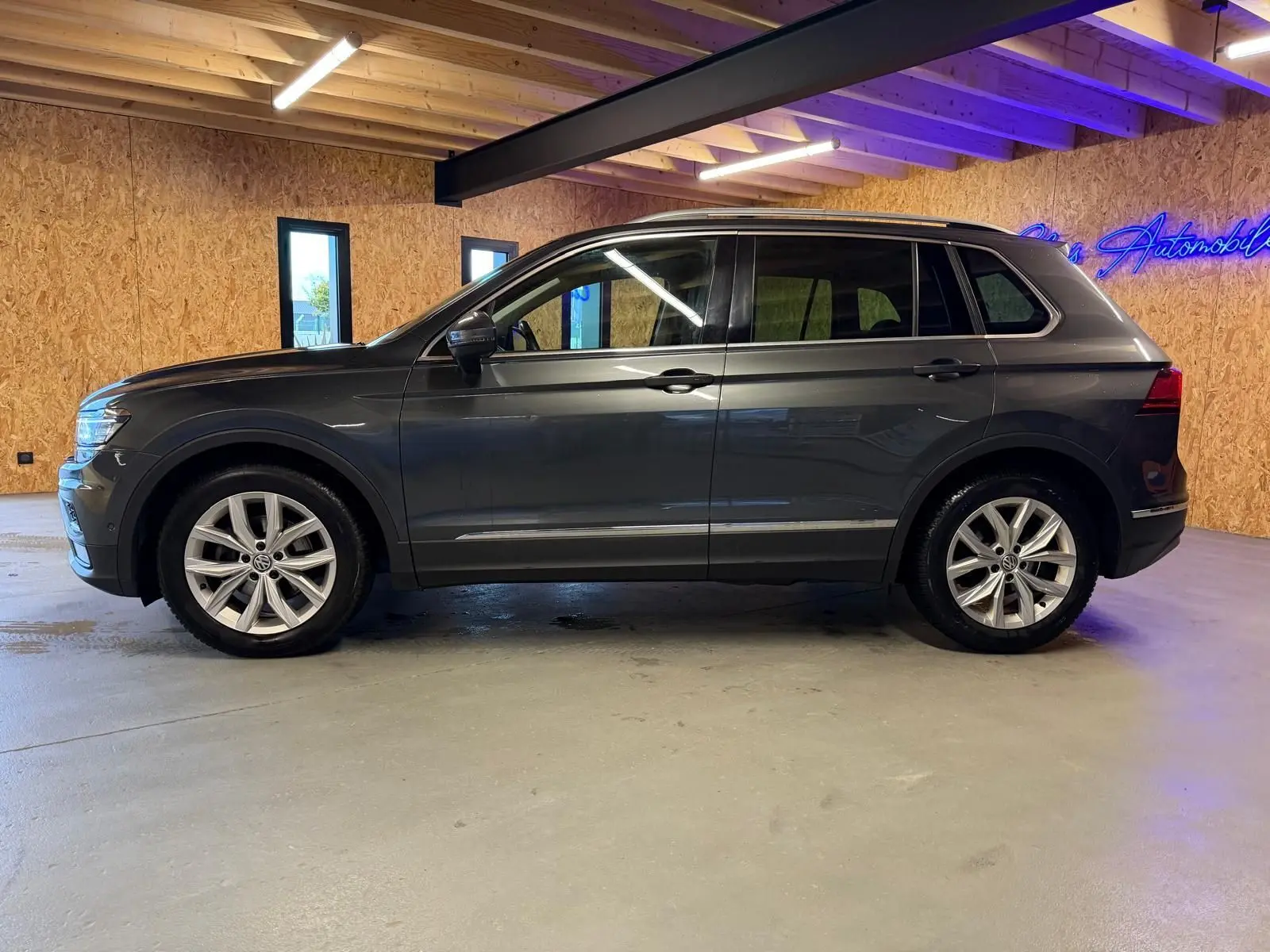 Vue de profil côté gauche d'un Volkswagen Tiguan gris avec jantes alu et barres de toit dans un garage.