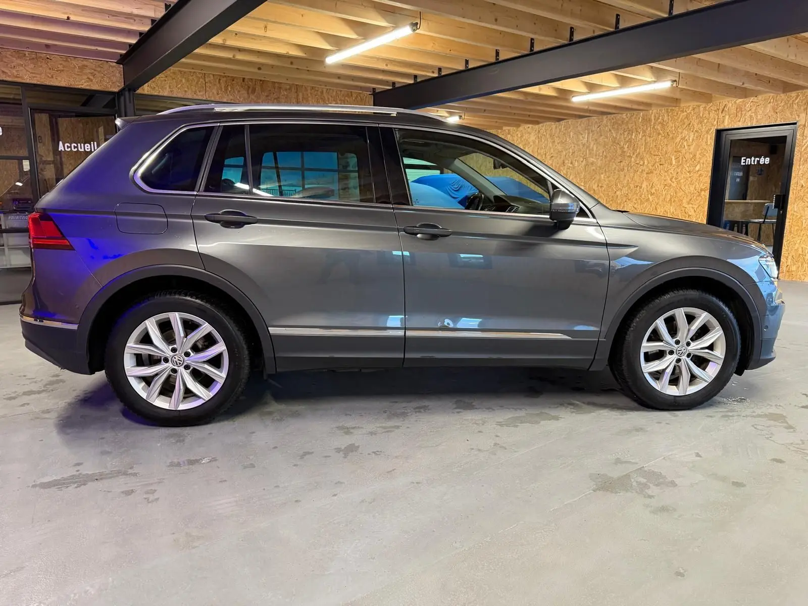 Vue de profil côté gauche d'un Volkswagen Tiguan gris avec jantes alu et barres de toit dans un garage.