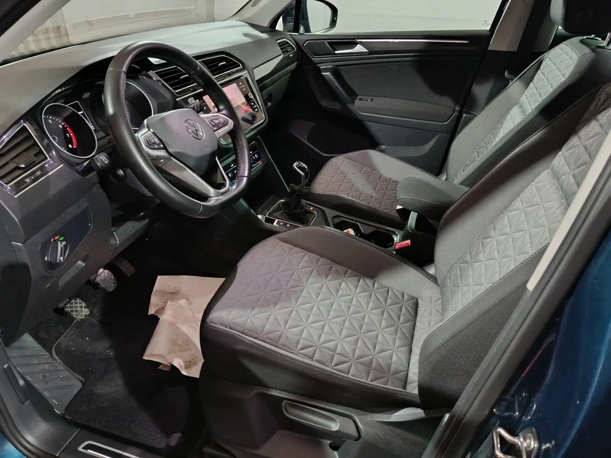Vue intérieure côté conducteur du Volkswagen Tiguan bleu, montrant le volant cuir multifonctions et les sièges avant gris à motifs.