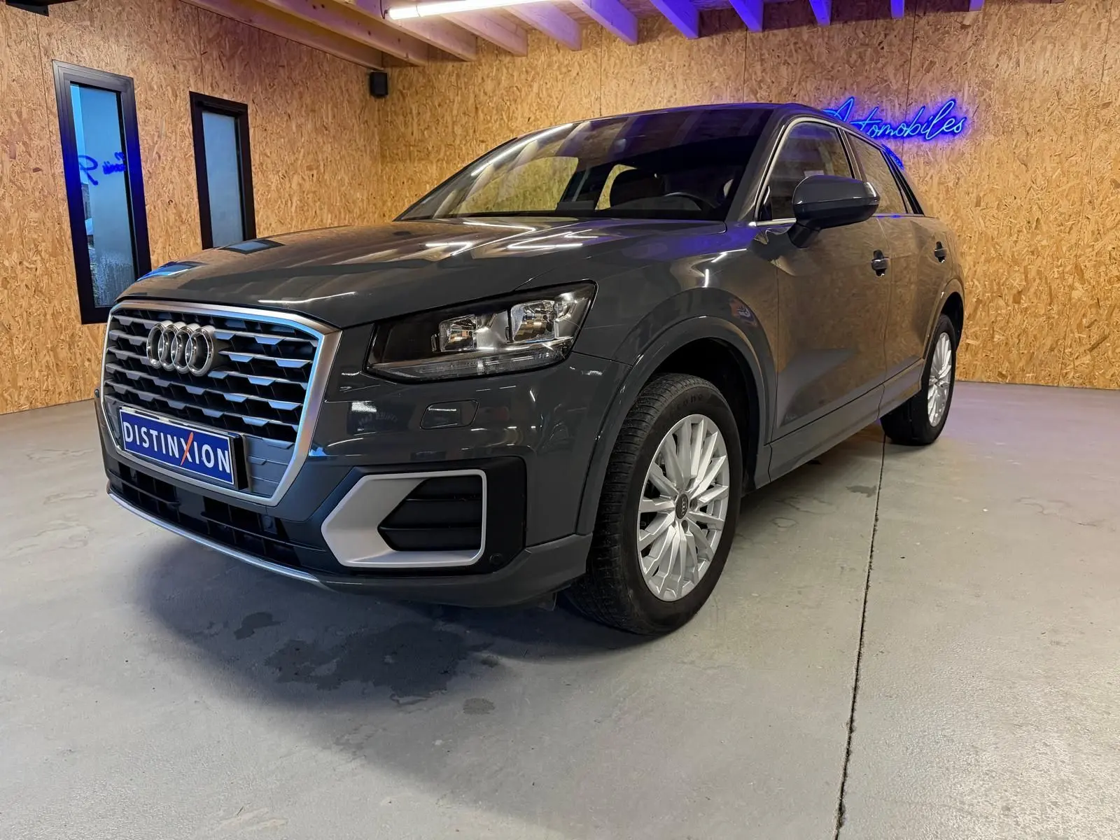 Audi Q2 gris en 3/4 avant droit dans un intérieur avec jantes alu et calandre chromée visible.
