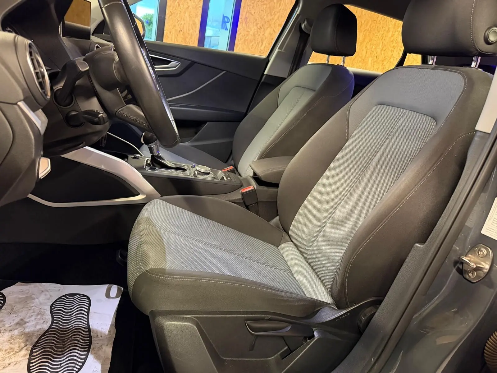 Vue intérieure côté conducteur de l'Audi Q2 gris, montrant les sièges bi-ton et la console centrale avec levier de vitesse automatique.