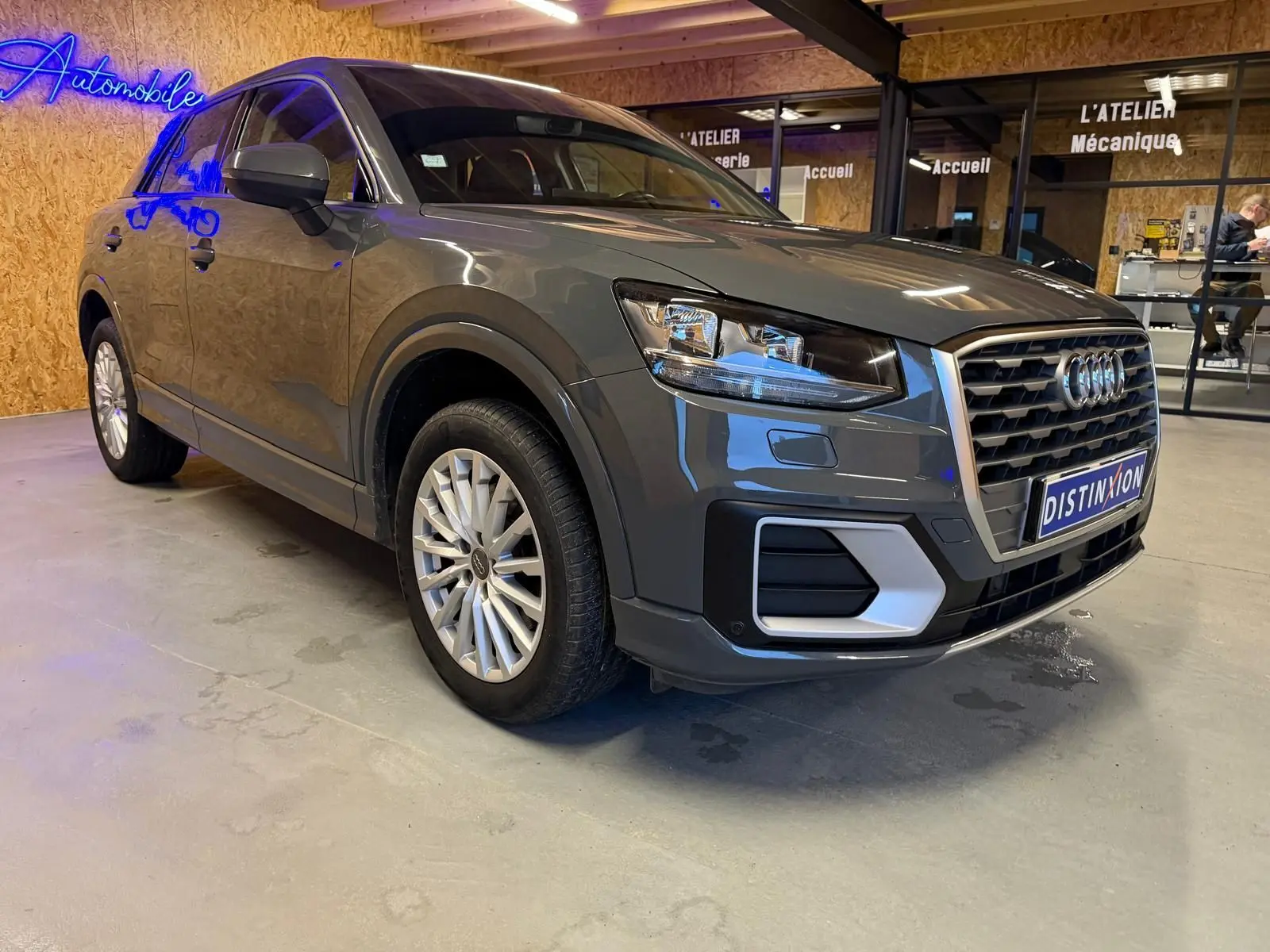 Audi Q2 gris vue 3/4 avant droit, jantes alu, calandre hexagonale et rétroviseurs électriques rabattables.