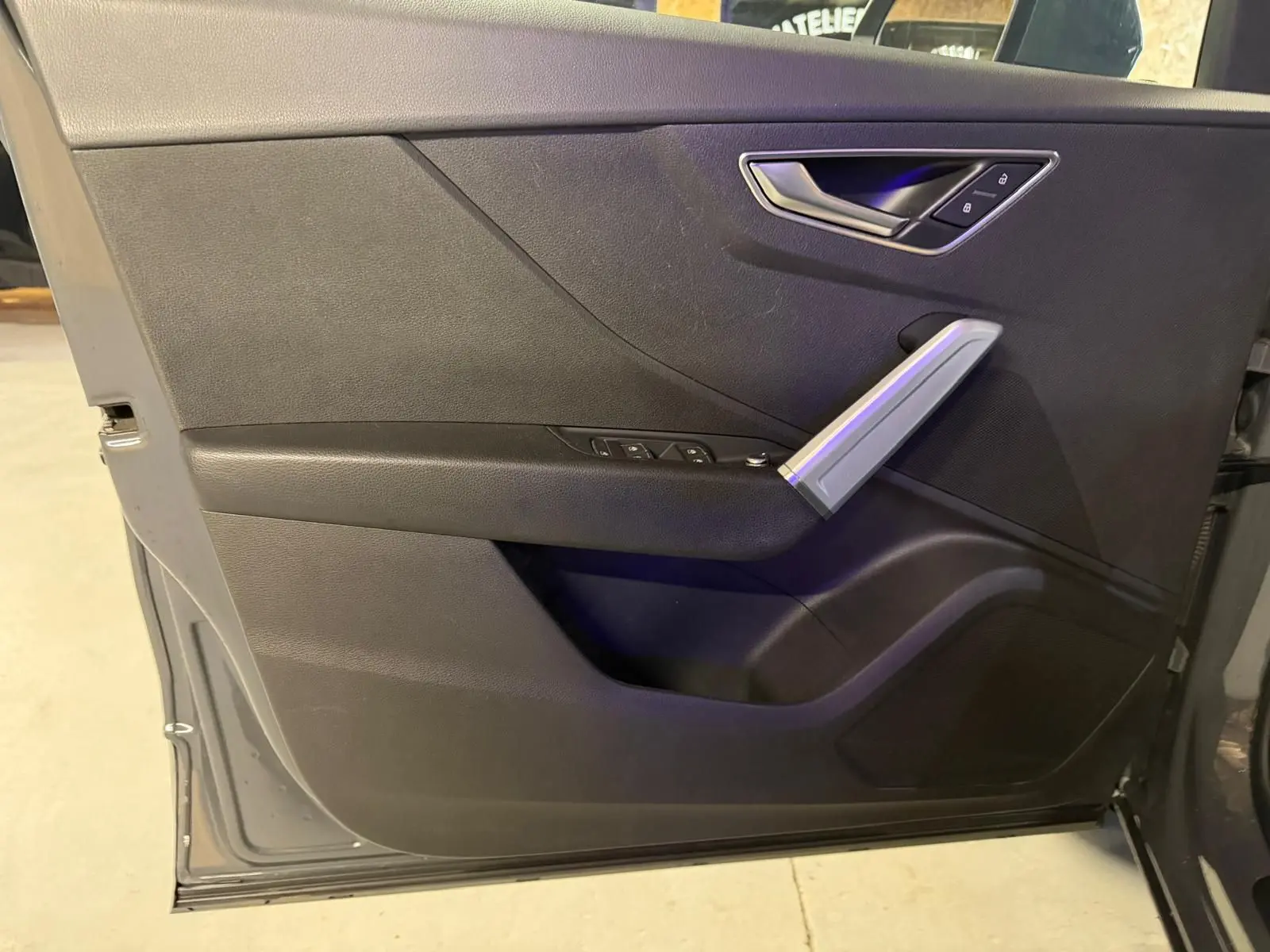 Vue rapprochée de la porte avant gauche de l'Audi Q2 gris, avec commandes électriques et poignée argentée.