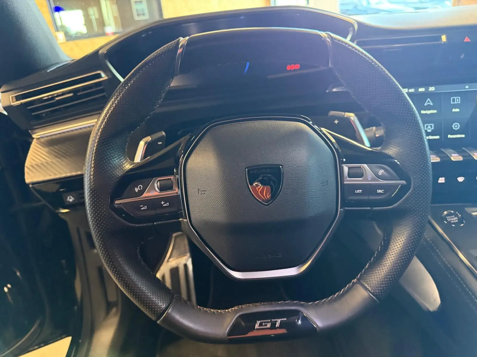 Volant cuir perforé noir avec badge Peugeot et sigle GT, tableau de bord et commandes tactiles en arrière-plan.