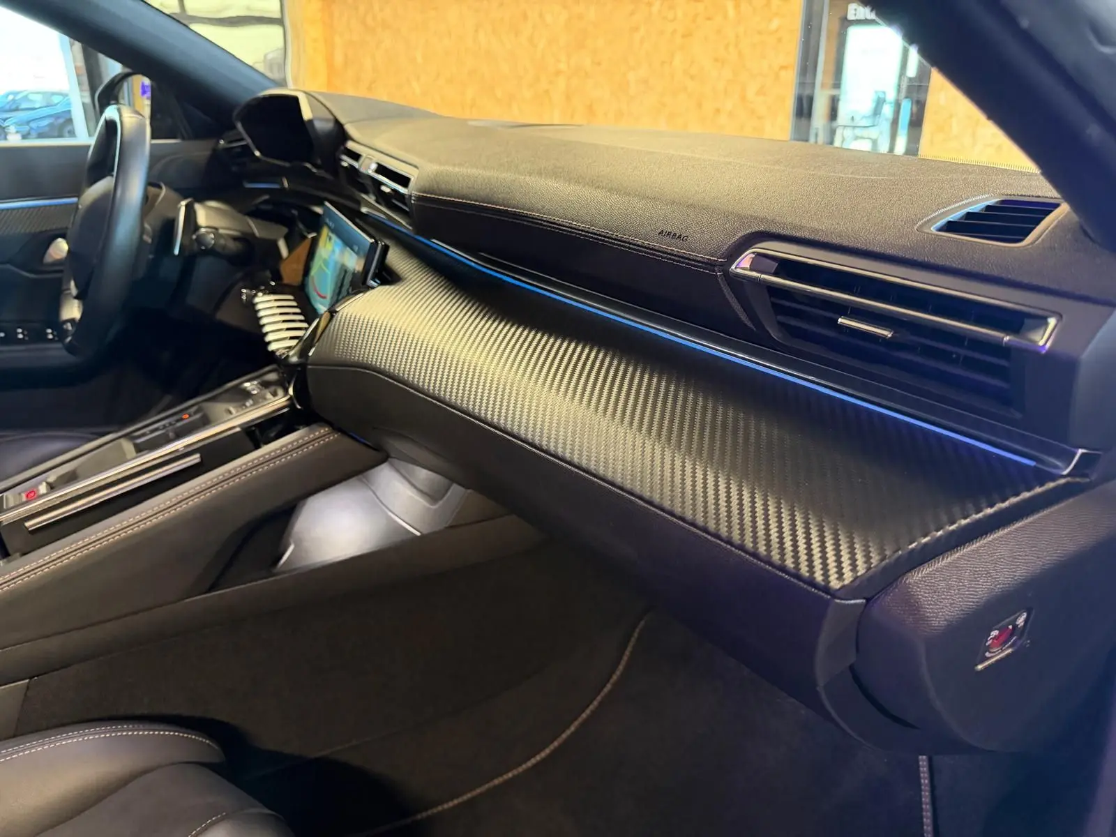 Vue intérieure côté passager du tableau de bord noir avec finition carbone et éclairage d'ambiance bleu sur Peugeot 508 SW 2024.