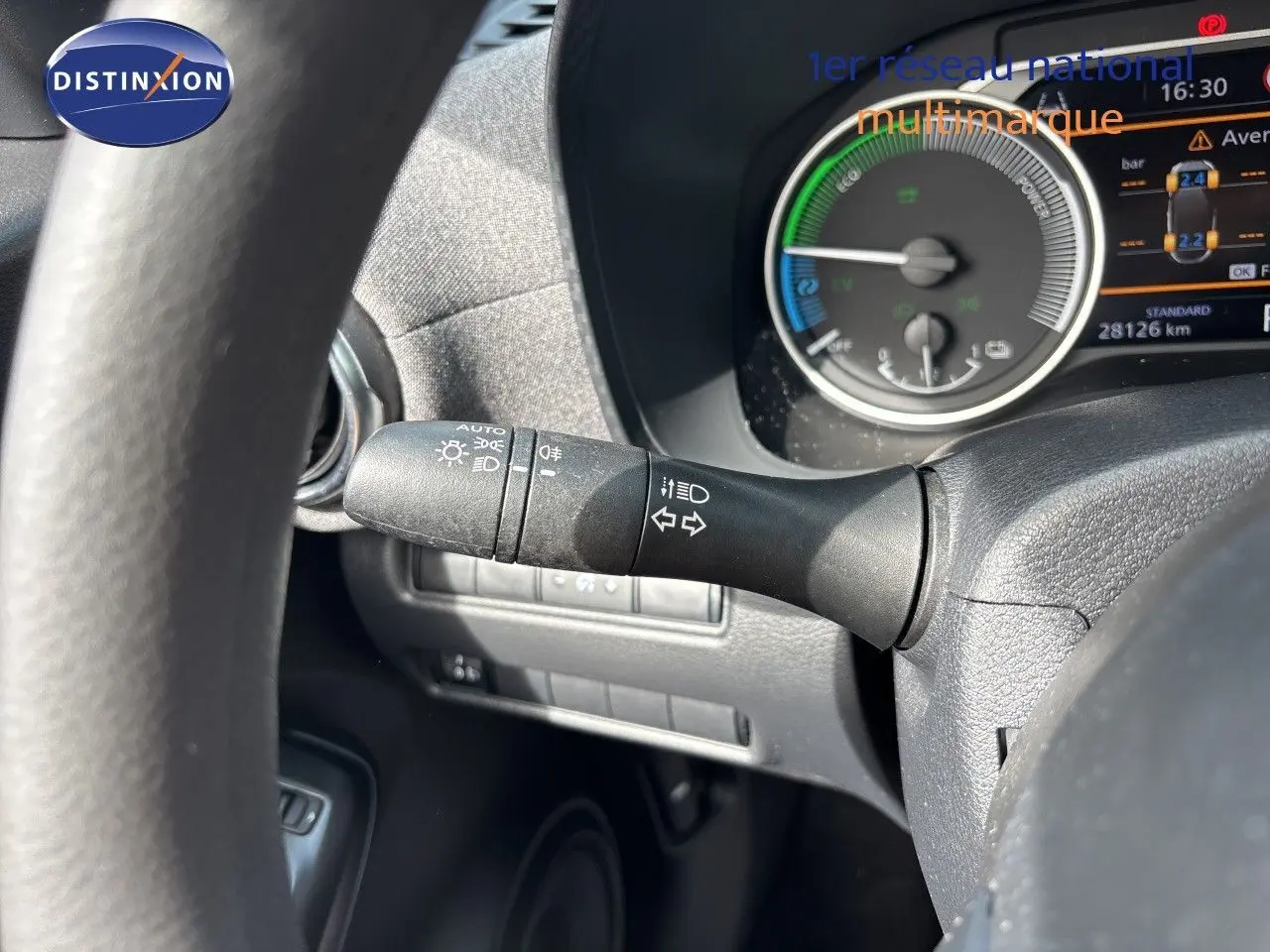 Gros plan sur le commodo d’éclairage à gauche du volant du Nissan Juke Hybrid gris, avec tableau de bord numérique en arrière-plan.