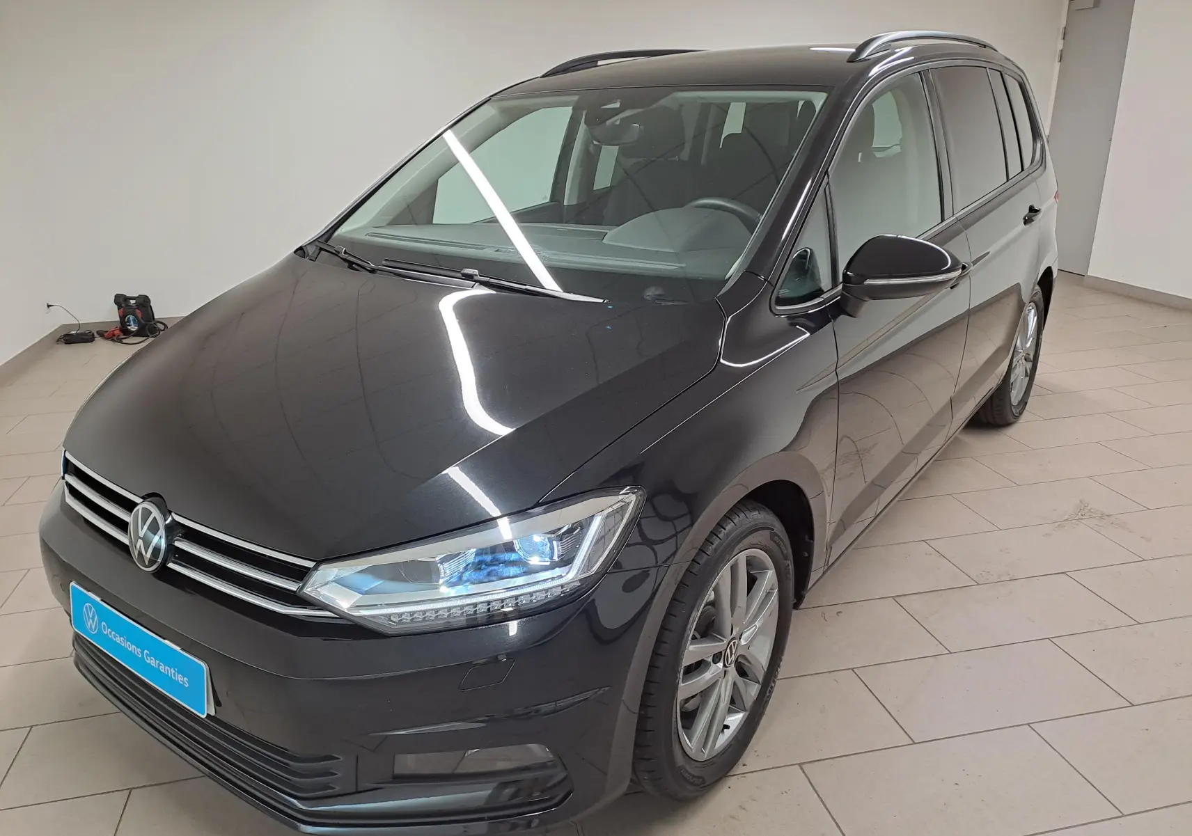 Volkswagen Touran noir grenadille vu en 3/4 avant droit dans un intérieur lumineux avec phares allumés.