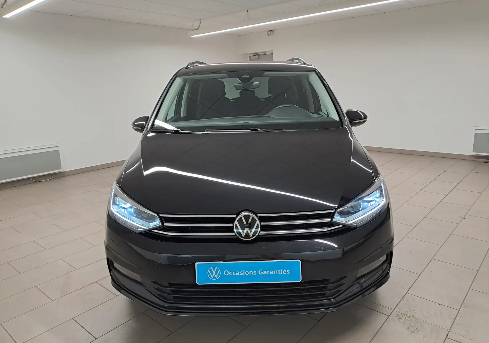 Vue avant d'un Volkswagen Touran 2025 noir grenadille avec phares allumés dans un showroom lumineux.