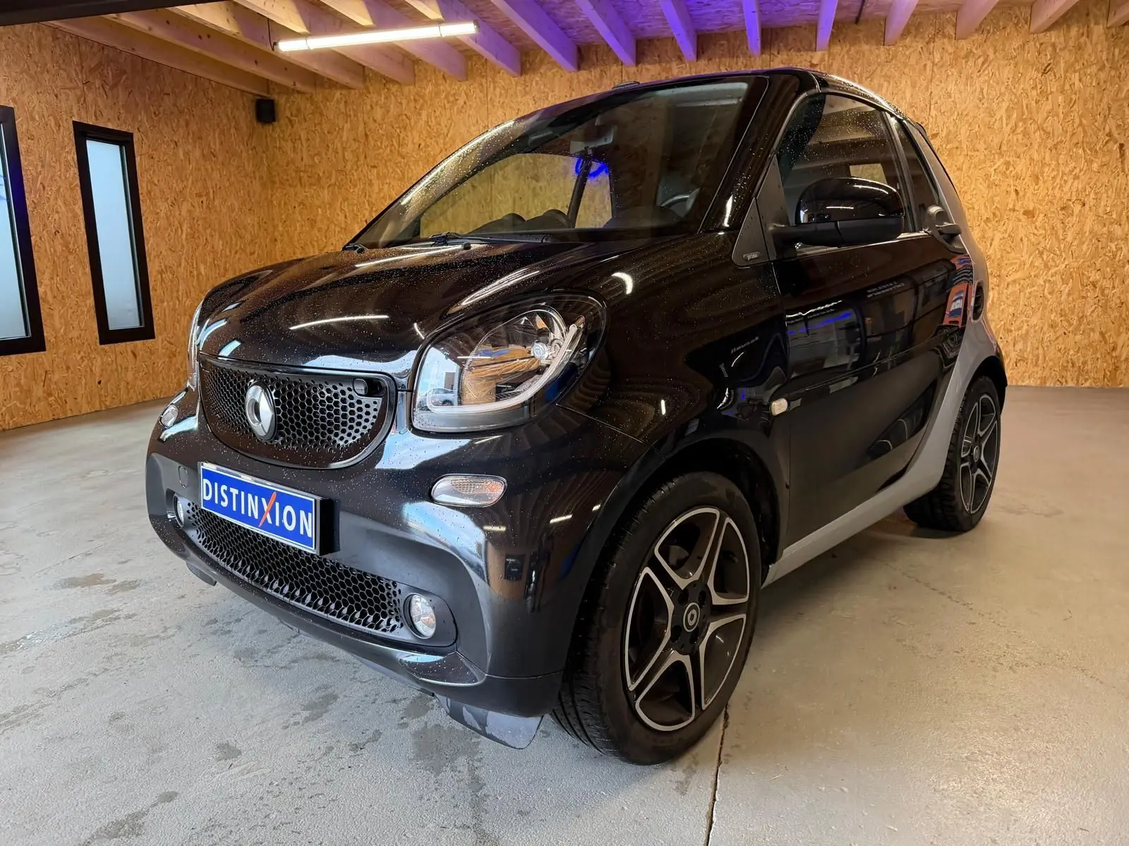 Vue 3/4 avant droite d'une Smart Fortwo Cabrio noire avec jantes alu 16 pouces dans un garage en bois.