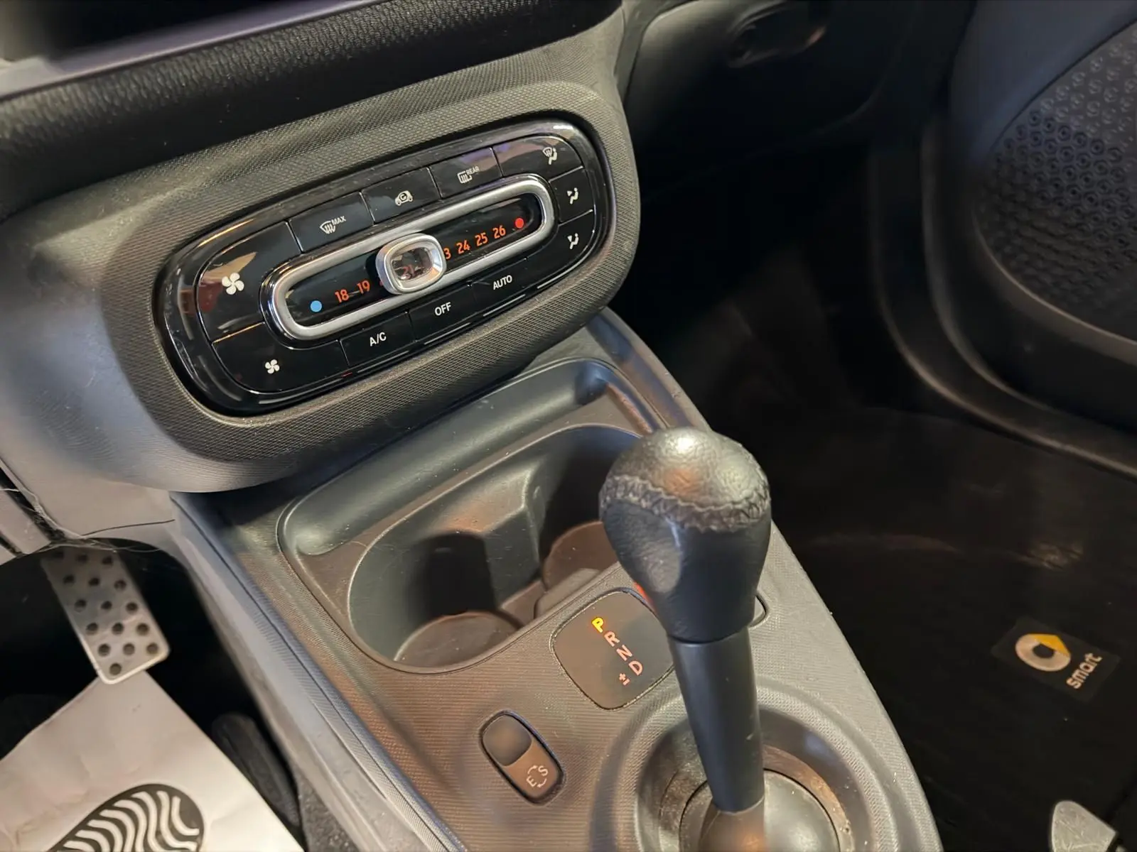 Gros plan intérieur sur la console centrale et levier de vitesse de la Smart Fortwo Cabrio noire, avec commandes climatisation et tapis logo Smart.