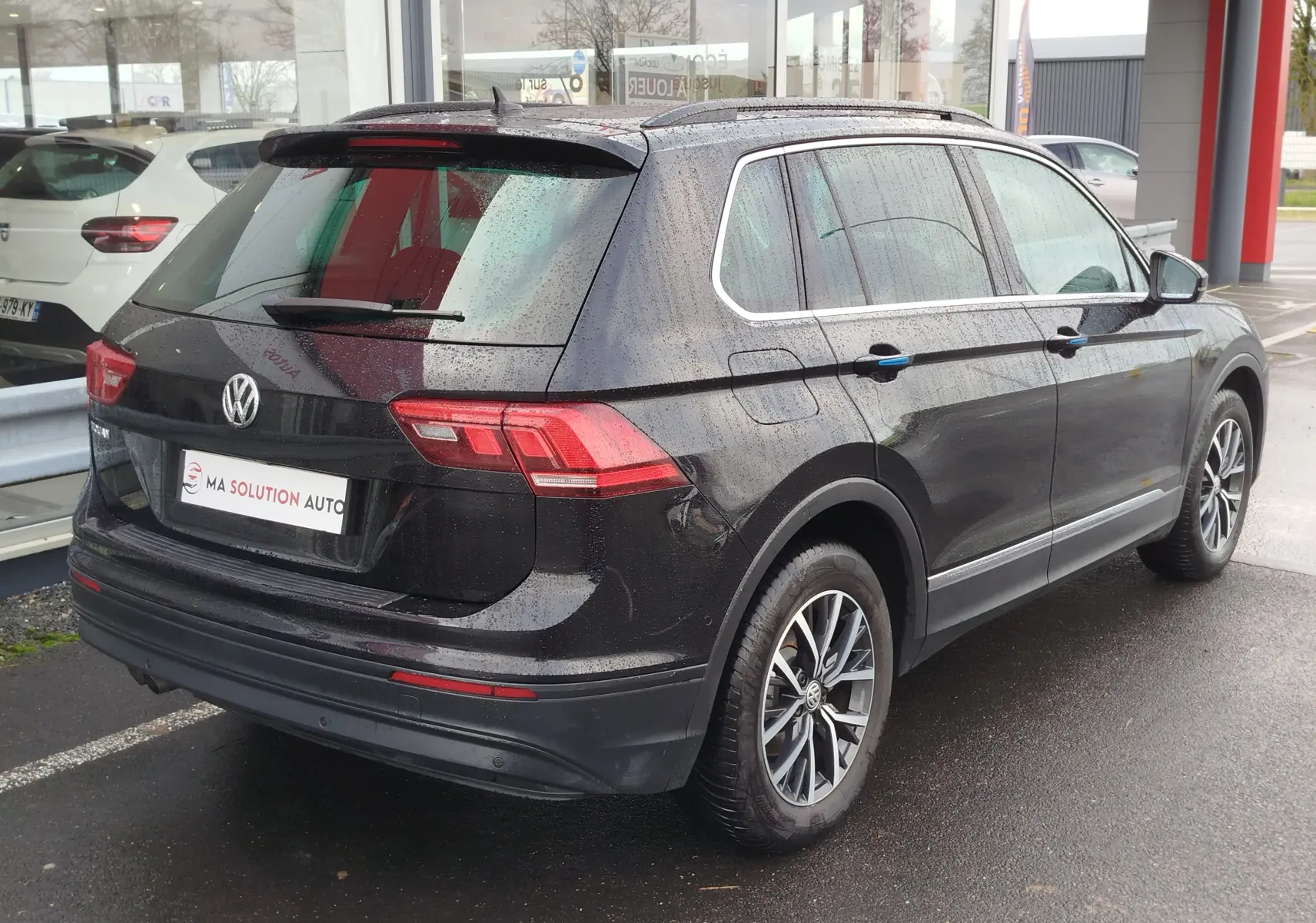 Volkswagen Tiguan noir intense nacré vu en 3/4 arrière droit sous la pluie, avec jantes alliage et vitres teintées.