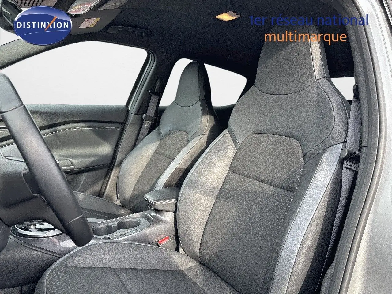 Intérieur du Nissan Juke 2024 montrant les sièges avant en tissu gris avec surpiqûres et accoudoir central.