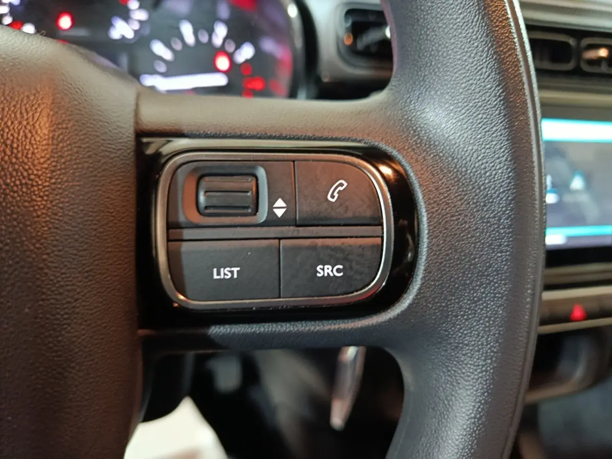 Gros plan sur les commandes gauche du volant multifonctions noir de la Citroën C3 2024, avec tableau de bord flou en arrière-plan.