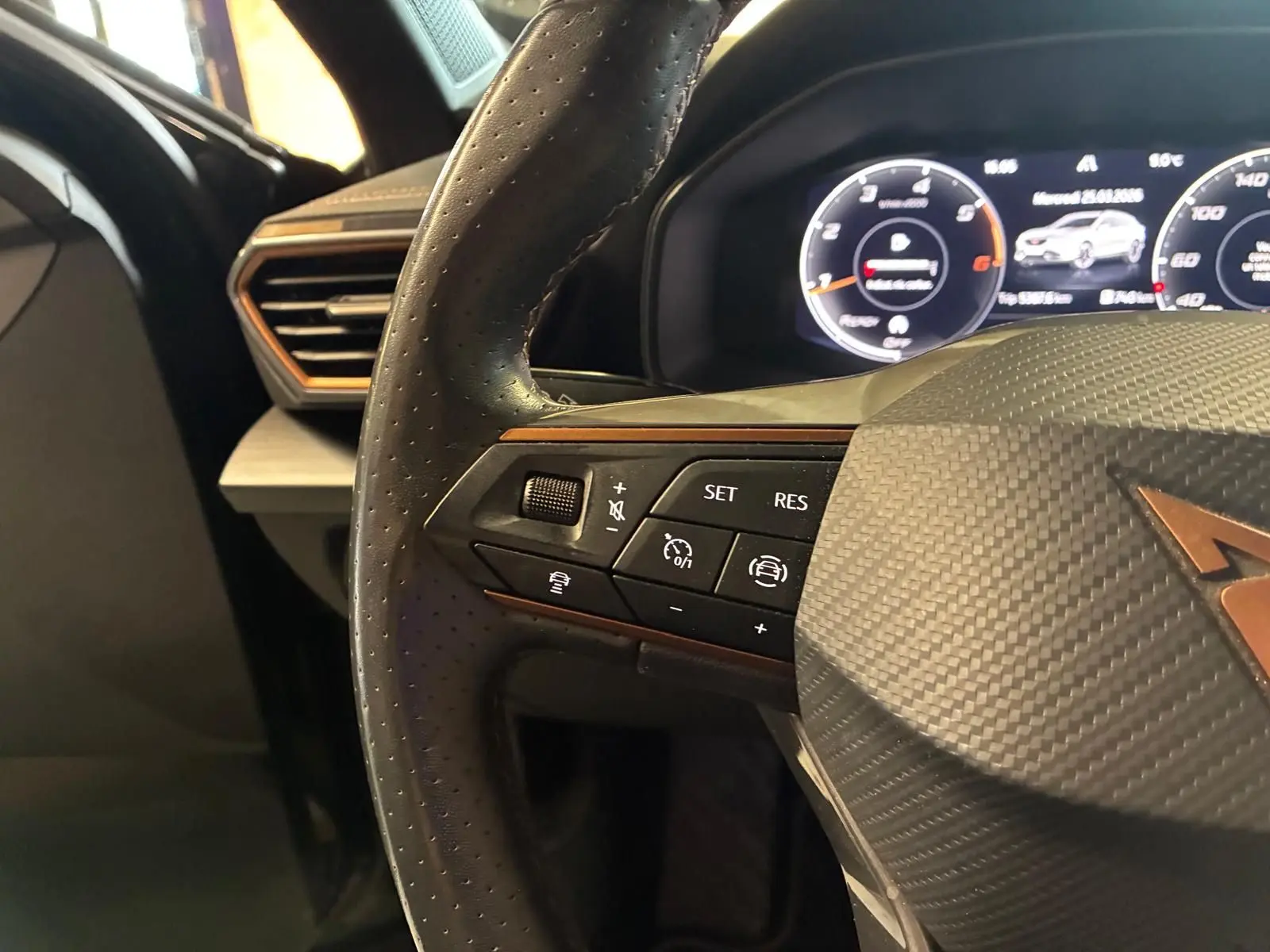 Gros plan sur le volant noir perforé avec logo CUPRA cuivré et commandes intégrées, tableau de bord numérique en arrière-plan.